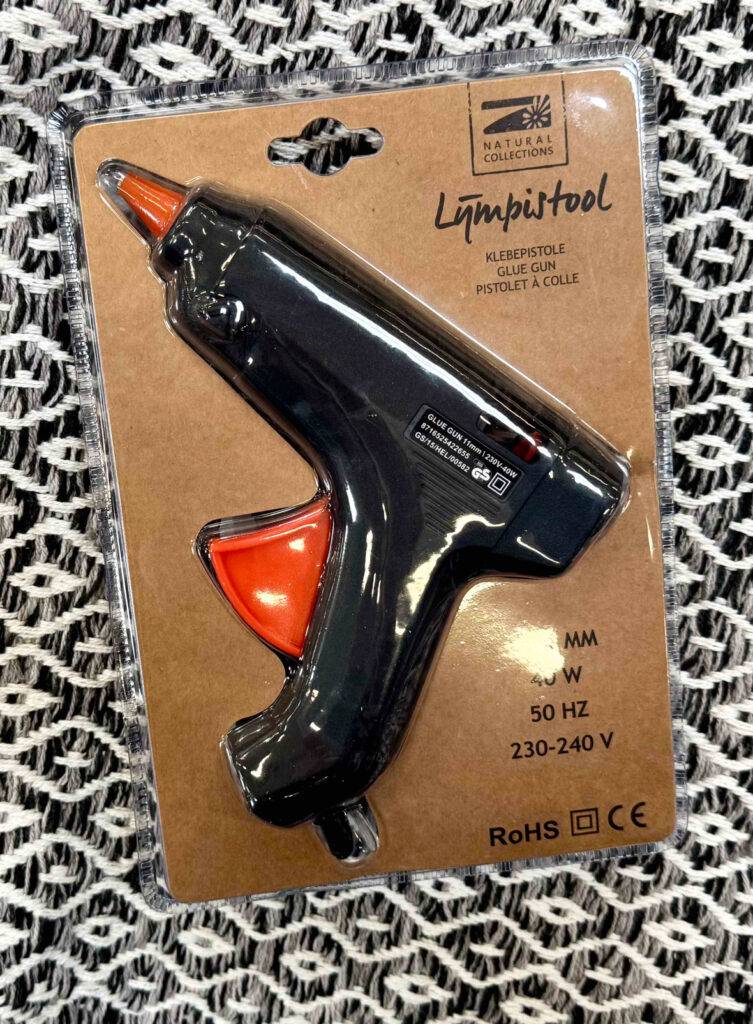 Die 11-mm-Heißklebepistole im Test liegt innerhalb der Verpackung auf einem gemusterten Stoff.