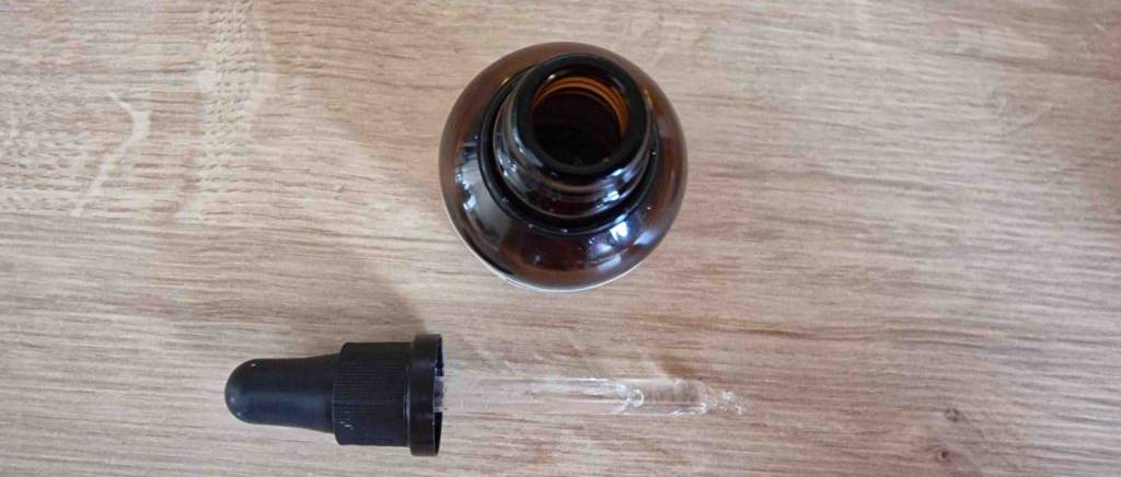 Ansicht von einem Vitamin-D-Präparat im Test in geöffneter, blickdichter Braunglas-Flasche neben seiner Pipette auf Holzuntergrund.