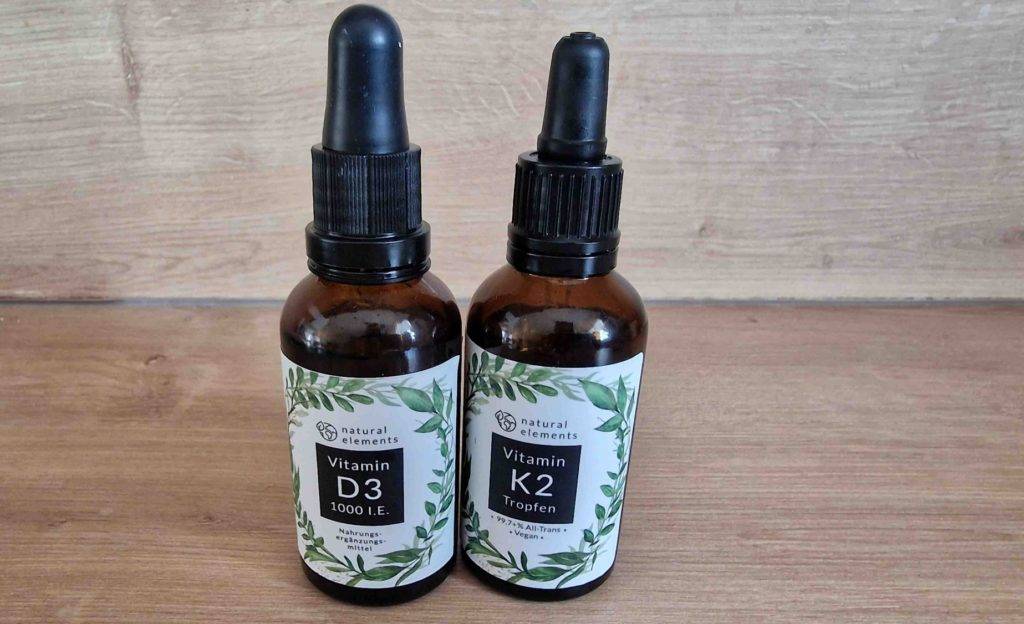 Natural Elements Vitamin-D-Präparat im Test befindet sich in verschlossener, blickdichter Braunglas-Flasche inklusive Pipette neben weiteren Tropfen mit Vitamin K2 vor einem Holzhintergrund.