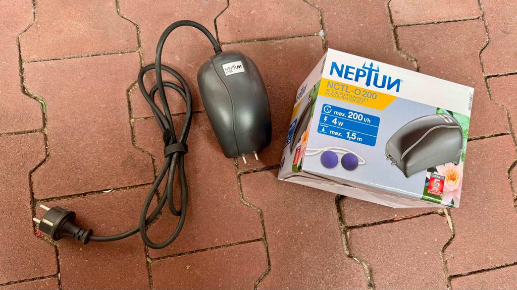 Ein Teichbelüfter liegt mit Stromkabel neben seinen Verpackungskarton zum Testen auf Betonfliesen.