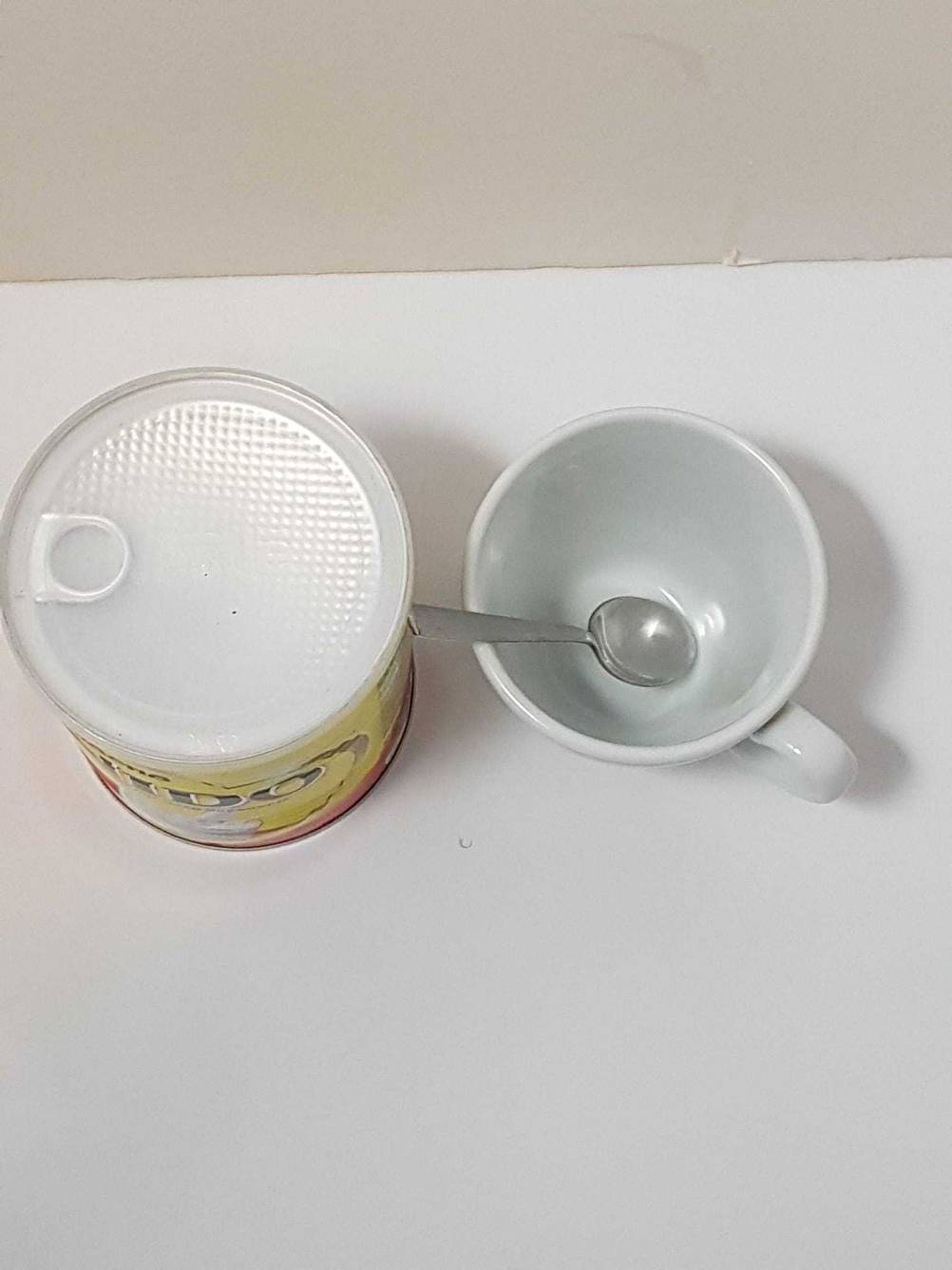 Milchpulver-Test: Eine Tasse neben einer Dose Milchpulver mit Löffel auf einer weißen Fläche stehend von oben.