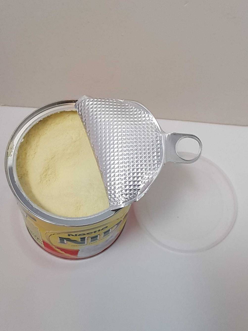 Eine getestete Dose Milchpulver mit geöffneter Versiegelung neben einem Deckel von oben auf einer hellen Fläche.