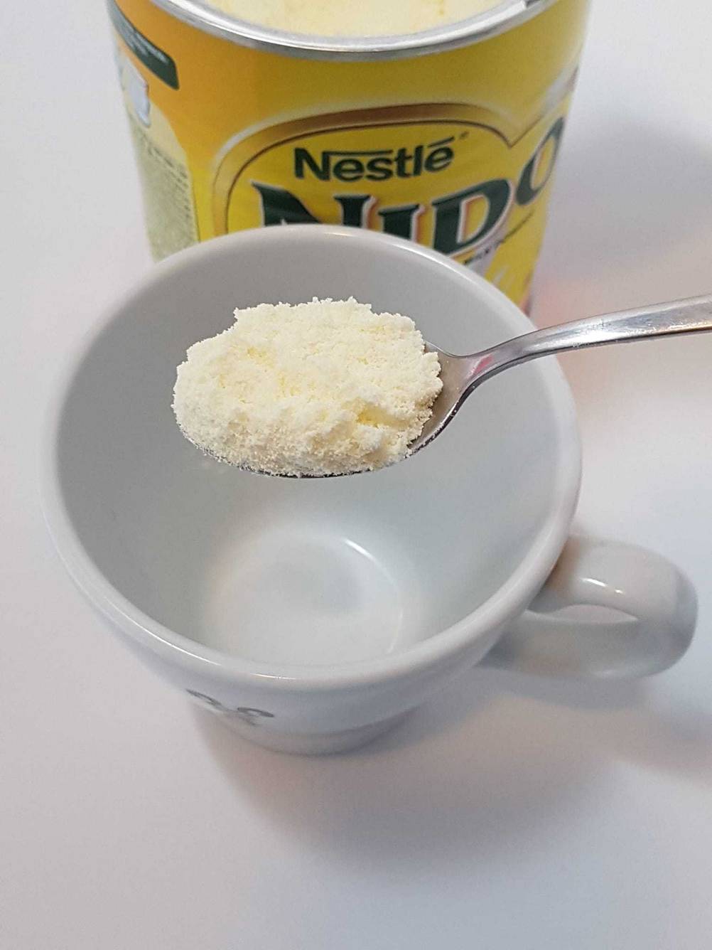 Ein gehäufter Löffel mit dem getesteten Milchpulver über einer Tasse, im Hintergrund eine Packung.