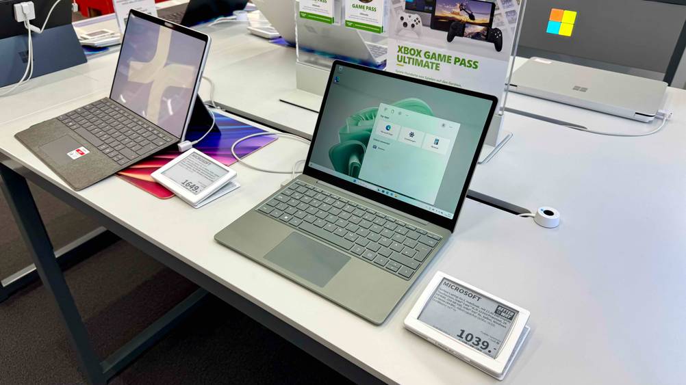 In einer Ausstellung befindet sich ein eingeschaltenes Netbook im Test mit Preisangabe neben anderen Geräten.