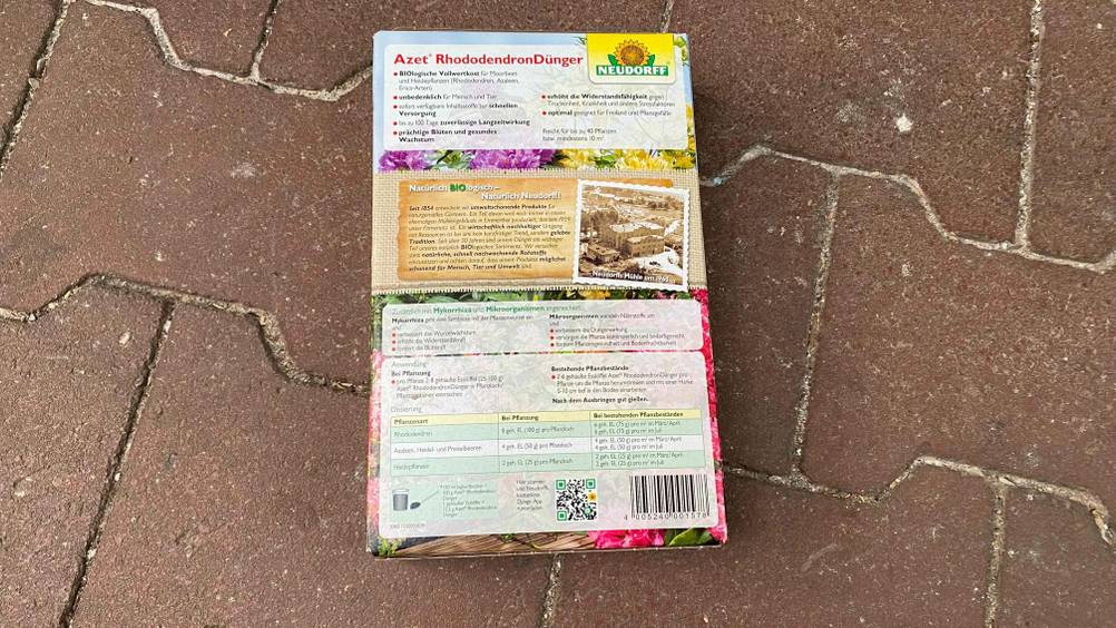 Heller unt bunter Karton mit Neudorff Rhododendron-Dünger im Test liegt mit Fokus auf die rückwärtige Produktbeschreibung auf Steinfliesen.