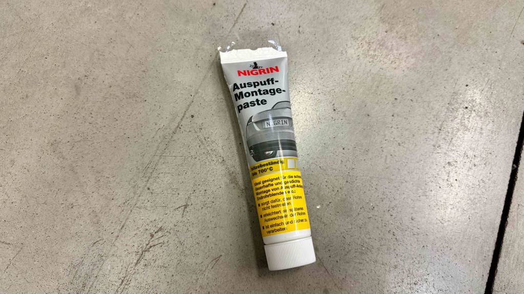 Eine Tube getestete Auspuff-Montagepaste liegt auf einem grauen Boden.