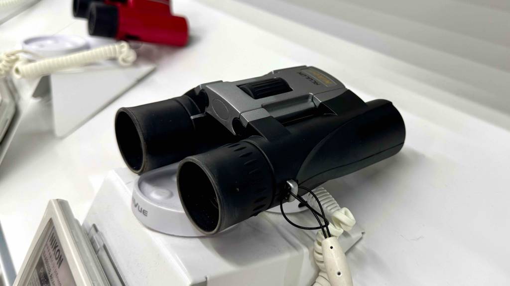 Test: 10 x 25-Ferngläser. Das schwarze Modell der Marke NIKON liegt gesichert vor einem roten Fernglas auf einer weißen Ausstellungsfläche.