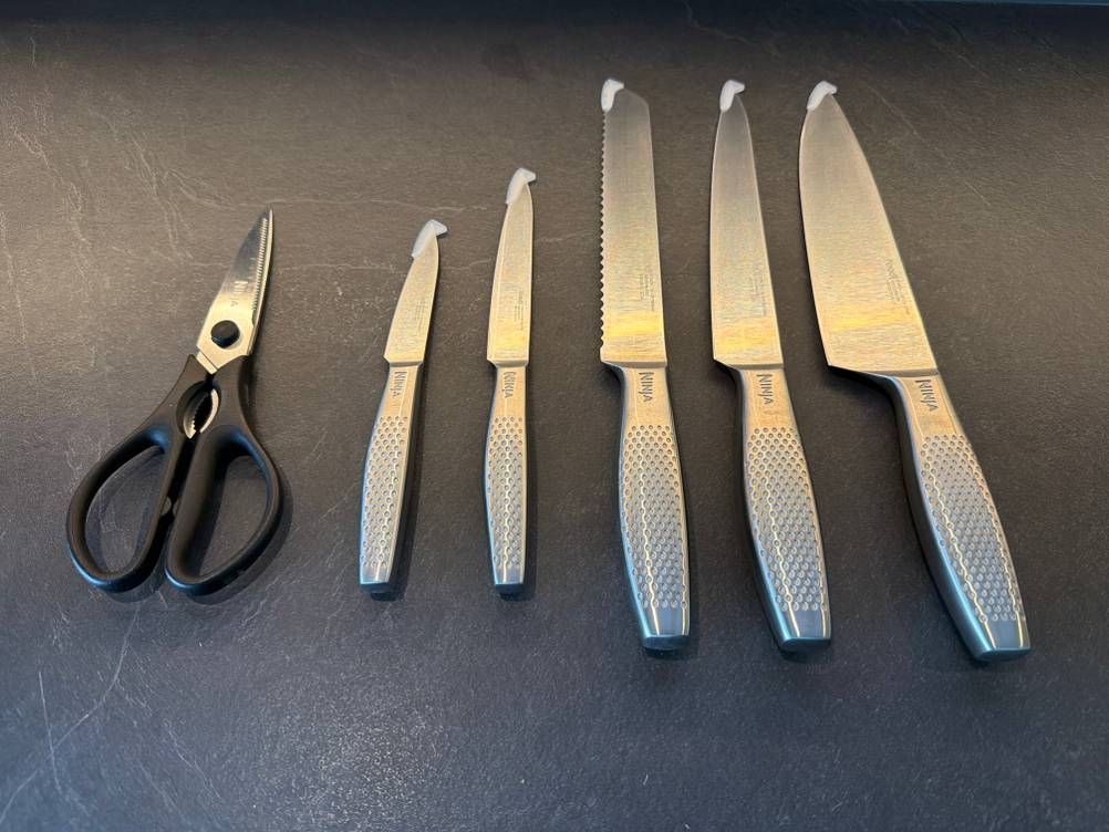 Fünf Messer und eine Schere liegen auf einer Küchenplatte.