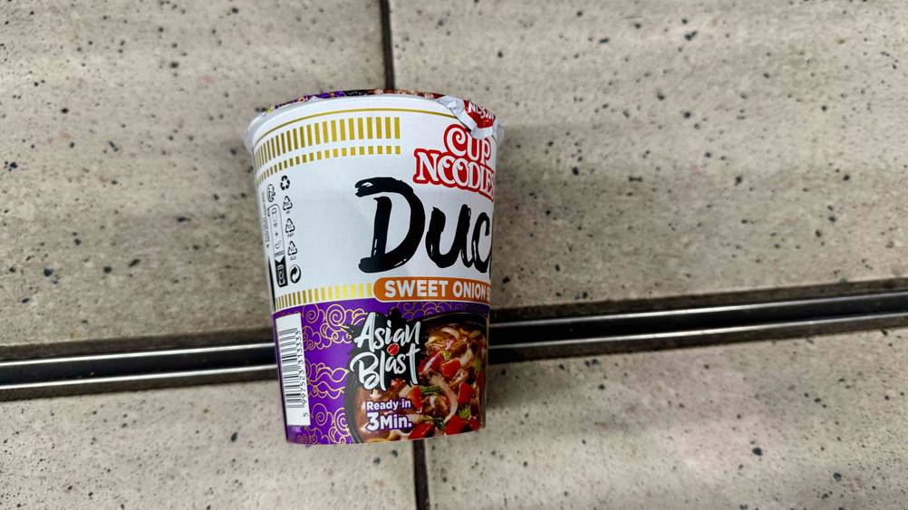 Die getesteten Nissin-Cup-Noodles seitlich auf einem Fließenboden liegend.