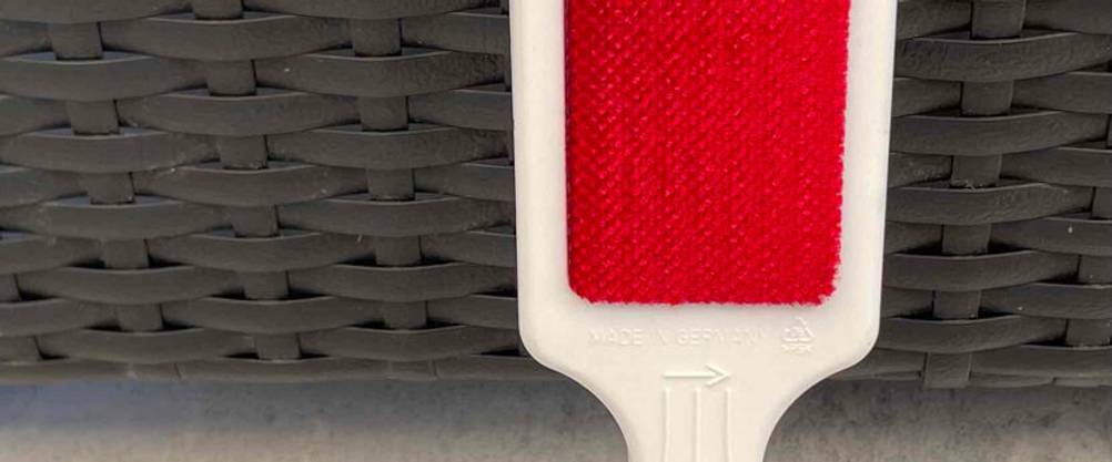 Eine weiße getestete Fusselbürste mit rotem reinigendem Velours-Material und richtungsweisendem Pfeil für die korrekte Anwendung liegt auf einer beigefarben Oberfläche.