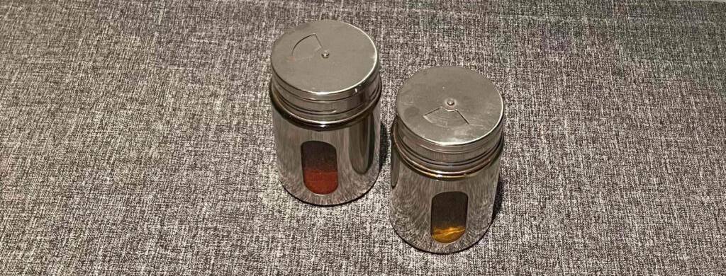 Zwei Aluminium-Gewürzstreuer inklusive Sichtfenster und Schraubverschluss mit Dosieröffnung, von schräg oben betrachtet, stehen auf einem grauen Stoff.
