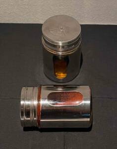Zwei getestete Gewürzstreuer aus Aluminium mit Sichtfenster inklusive rotem und orangem Pulver befinden sich von schräg oben betrachtet auf einer schwarzen Oberfläche.