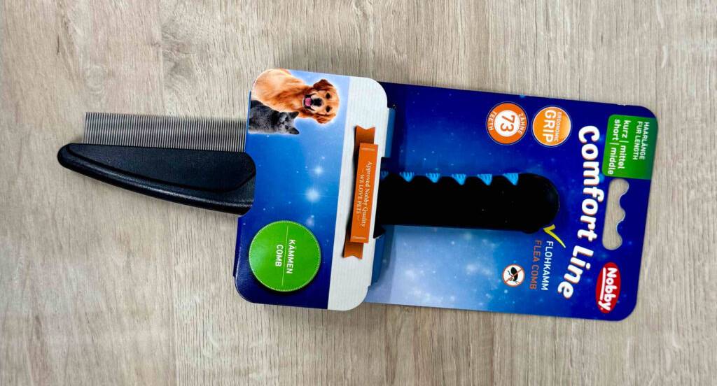 Der getestete Hundekamm in Schwarz liegt in der blauen Packung auf einem Holzboden.