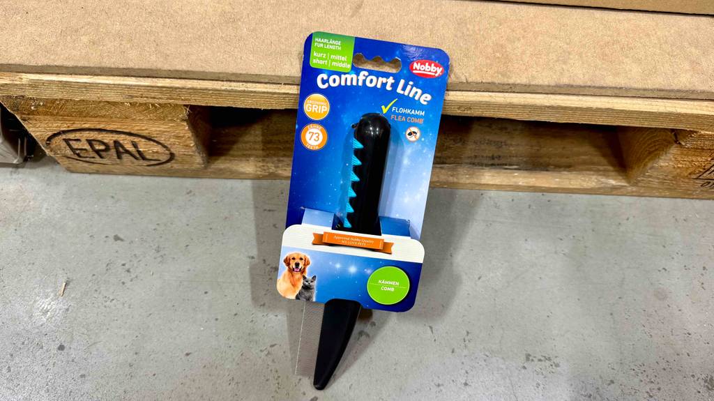 Auf einer Holzpalette liegt der getestete Hundekamm in Schwarz in der blauen Packung.
