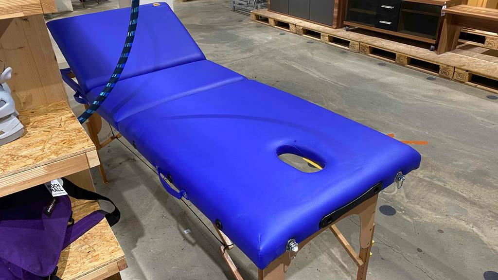Eine tragbare blaue Massageliege im Test, mit Fokus auf die Materialbeschaffenheit befindet sich mit aufgeklapptem Gestell neben einem Verkaufsregal.