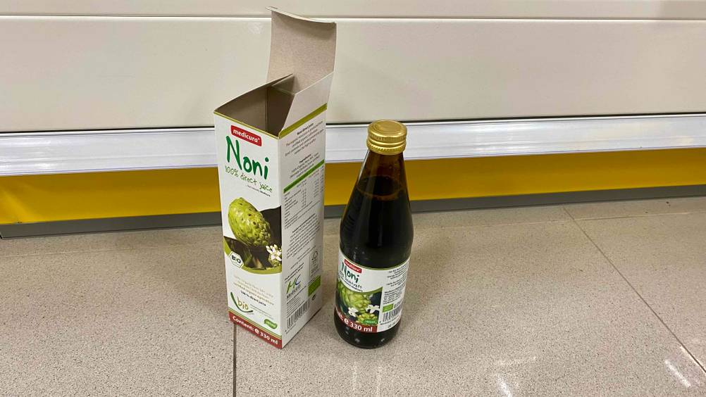 Neben der geöffneten Verpackung steht eine Flasche des getesteten Noni-Safts auf einem Fließenboden von schräg oben.
