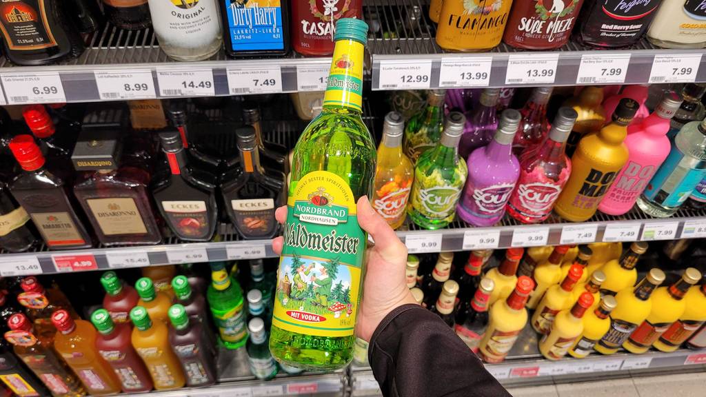 Eine Hand hält eine Flasche Waldmeisterlikör zum testen vor einem Spirituosenregal.