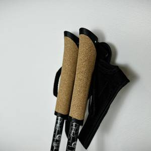 Zwei Alpidex-Nordic-Walking-Stöcke mit Korkgriffen und befestigten Handschlaufen lehnen an einer weißen Wand. Die Griffe sind nach oben ausgerichtet.