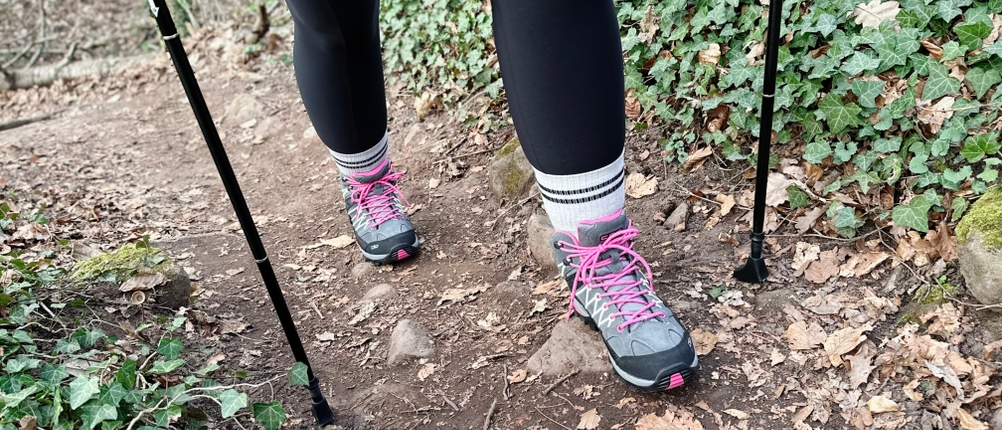 Eine Person steigt mit Glymnis-Walking-Stöcken einen bewachsenen Hang hinauf. Der Fokus liegt auf den Füßen und den Stöcken.