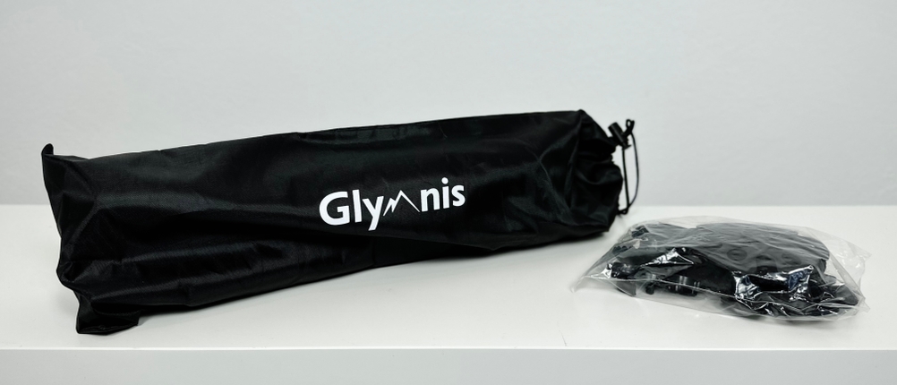 Eine schwarze Glymnis-Tasche mit Logo liegt auf einem Tisch. Daneben befindet sich das Zubehör in einem transparenten Plastikbeutel.