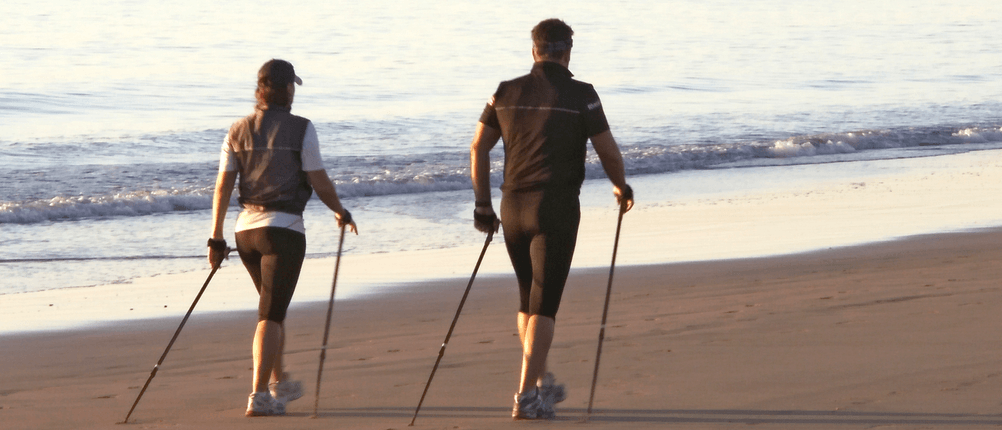 Zwei Personen laufen mit Nordic-Walking-Stöcken am Strand entlang. Sie sind nur von hinten zu sehen.