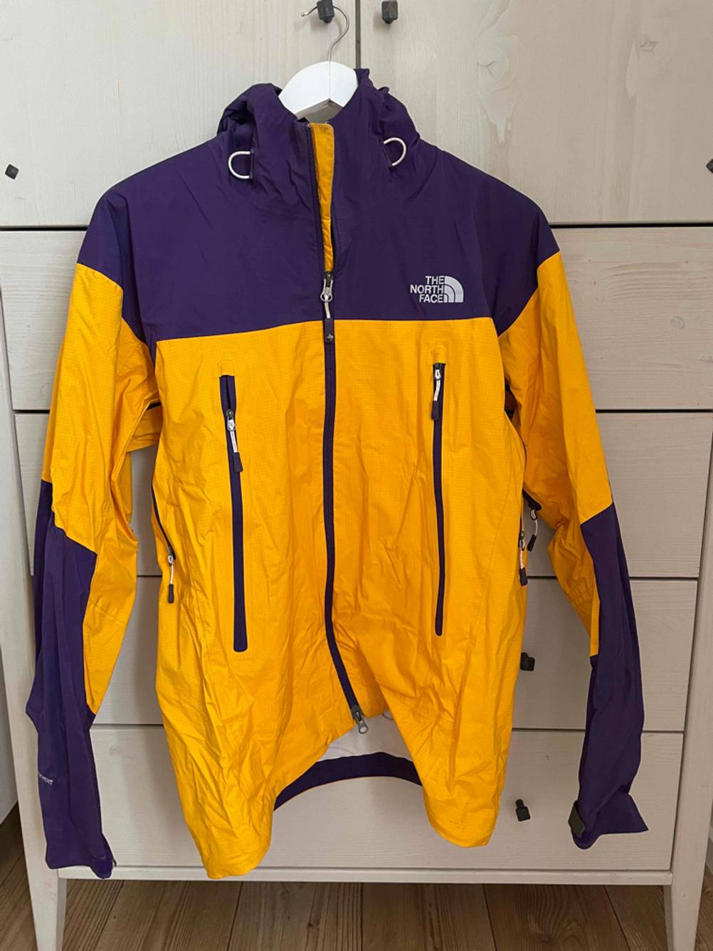 Regenjacke-Unisex-Test: Frontansicht einer getesteten lila-gelben Unisex-Regenjacke, die vor einem Regal hängt.
