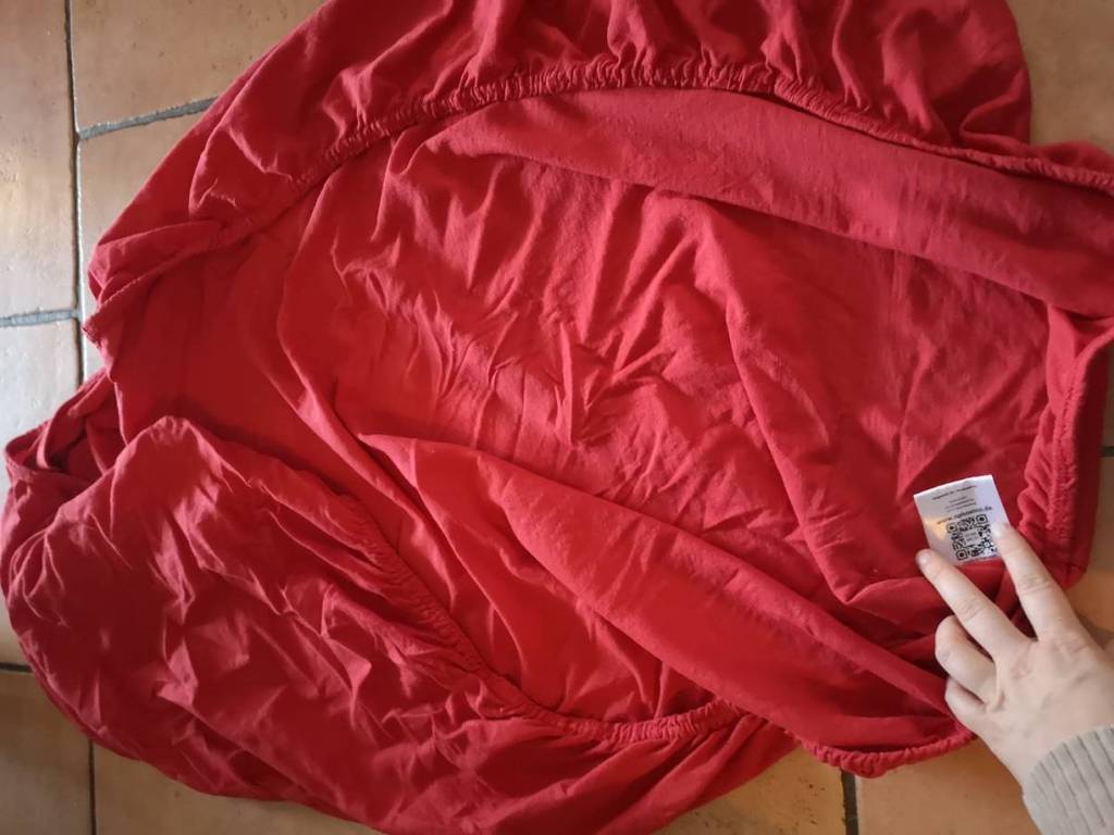 Hand zeigt auf ein weißes Produktetikett von einem roten getesteten Spannbettlaken, das ausgebreitet auf dem Boden liegt.