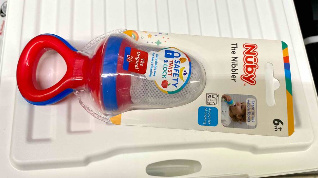 Ein getesteter Fruchtsauger von Nuby im blau-roten Design liegt innerhalb seiner transparenten Verpackung auf einem weißen Tablett.
