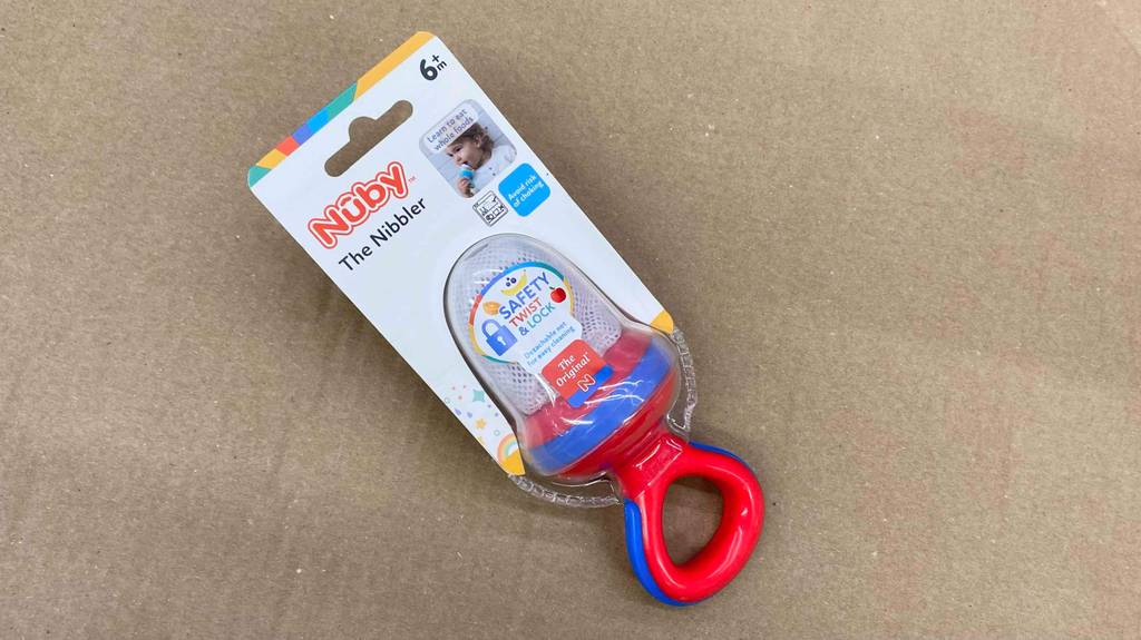 Ein rot-blauer Nuby Fruchtsauger im Test befindet sich in seiner transparenten Verpackung auf einem braunen Karton.