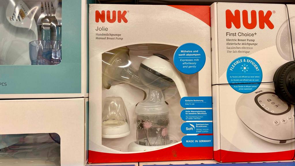 Eine Verpackung mit Sichtfenster, hinter dem sich eine getestete Hand-Milchpumpe der Marke NUK inklusive Zubehör befindet, steht neben einer elektrischen Milchpumpe von NUK.