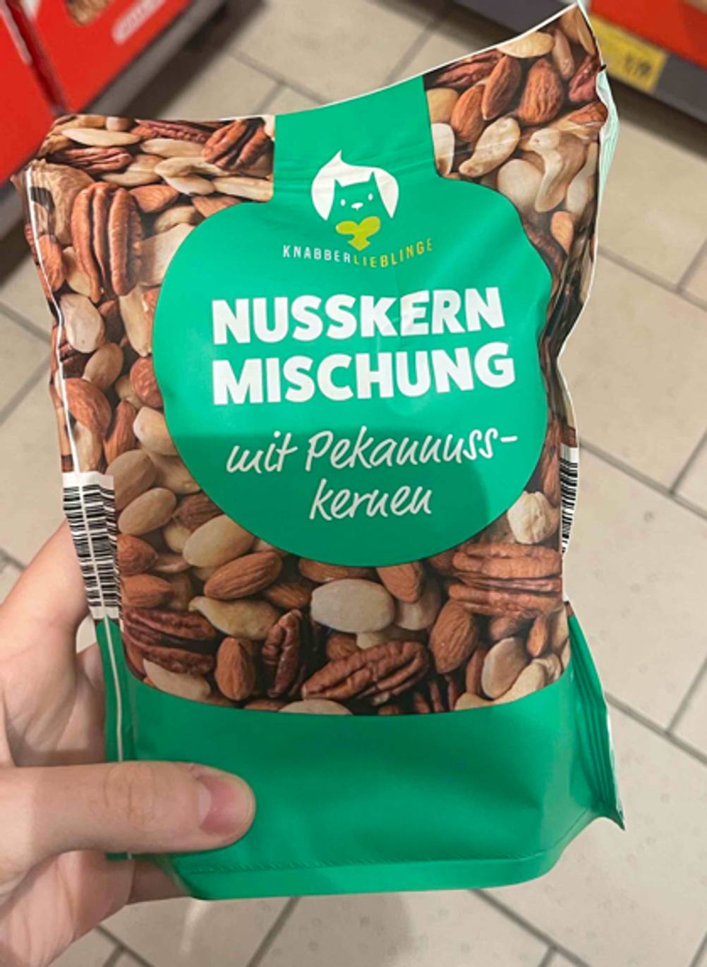 Im Test: Die Nussmischung mit Peannuss-Kernen in einer wiederverschließbaren Tüte im grünen Design wird von einer Hand über einem weißen Fliesebelag gehalten.
