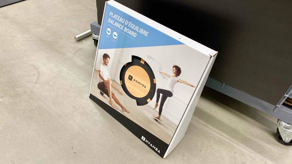 Ein originalverpacktes getestetes Balance-Board lehnt an einer dunklen Fläche.