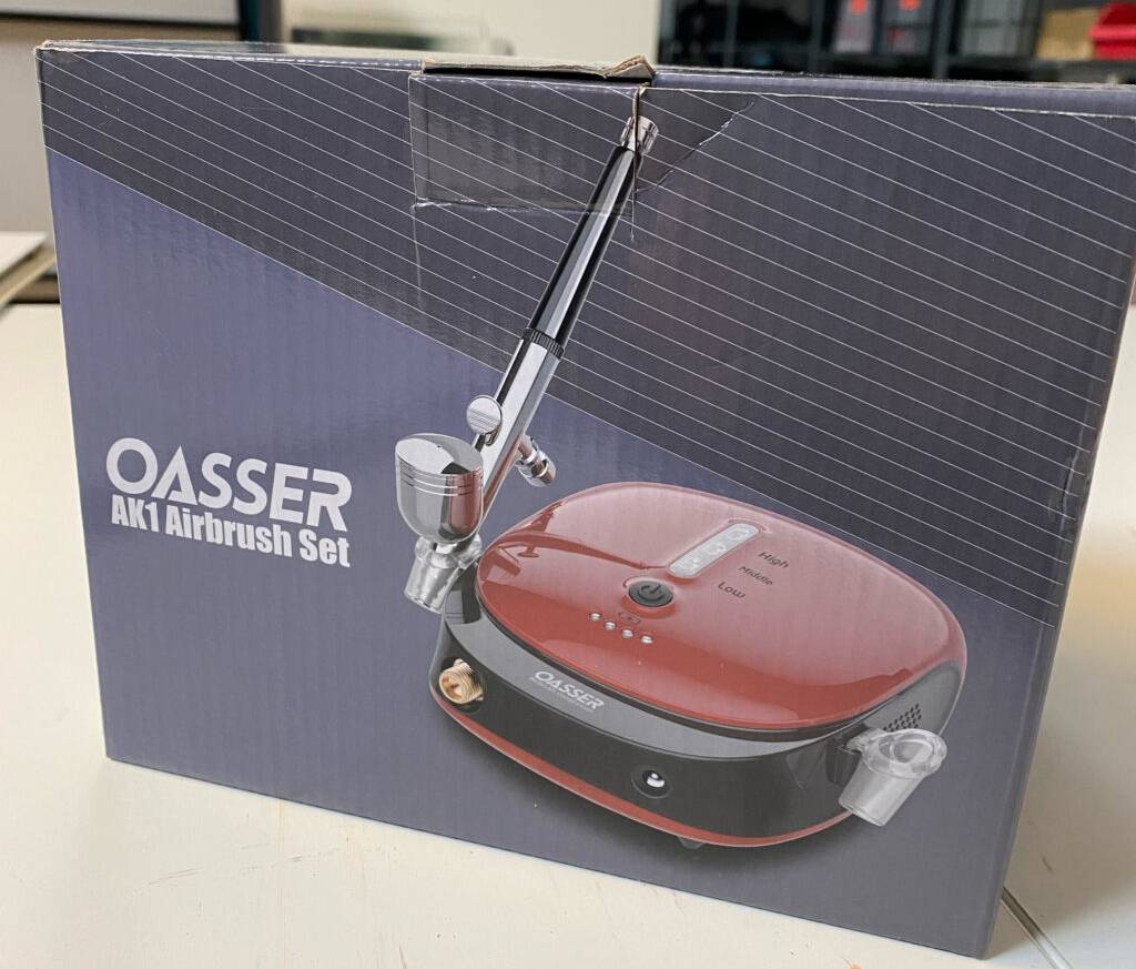 Eine Verpackung inklusive getestetem Airbrush-Set der Marke OASSER steht auf einem weißen Tisch.