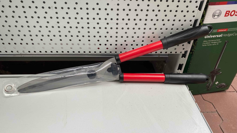 Eine getestete Heckenschere lehnt auf einem hellen Regalboden an einem Lochblech.