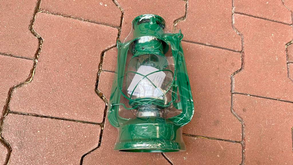 Eine grüne, in Zellophan verpackte und getestete Petroleumlampe befindet sich auf einem rötlichen Untergrund.
