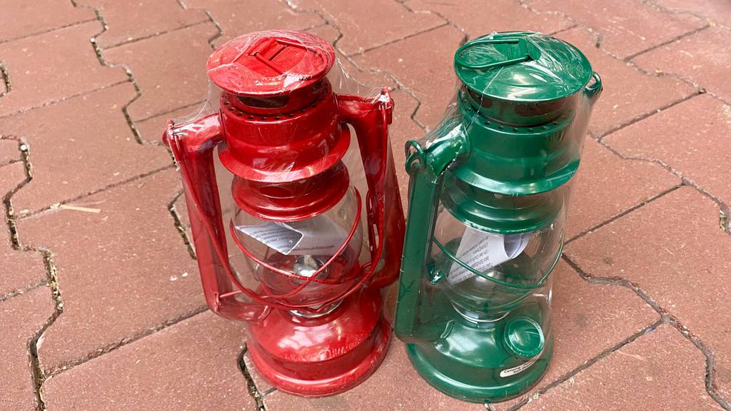 Petroleumlampe im Test: Zwei klassische Modelle, ein rotes und ein grünes, stehen in Zellophan verpackt auf einem Terracottaboden.