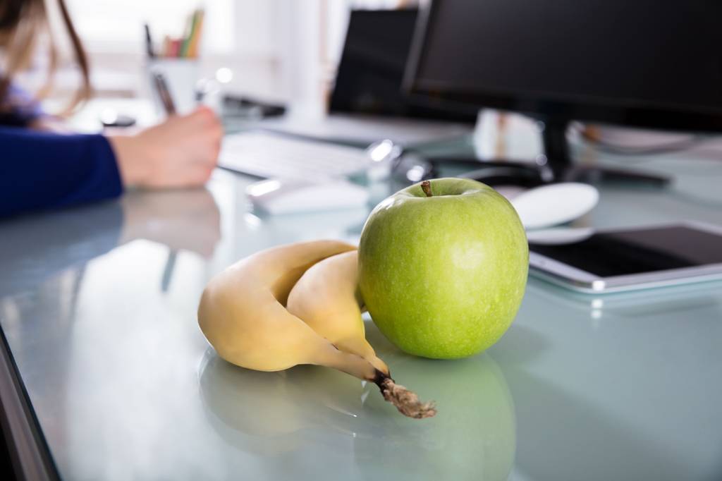 Zwei Bananen und ein Apfel liegen auf einem Schreibtisch im Büro. Im Hintergrund arbeitet eine Frau.