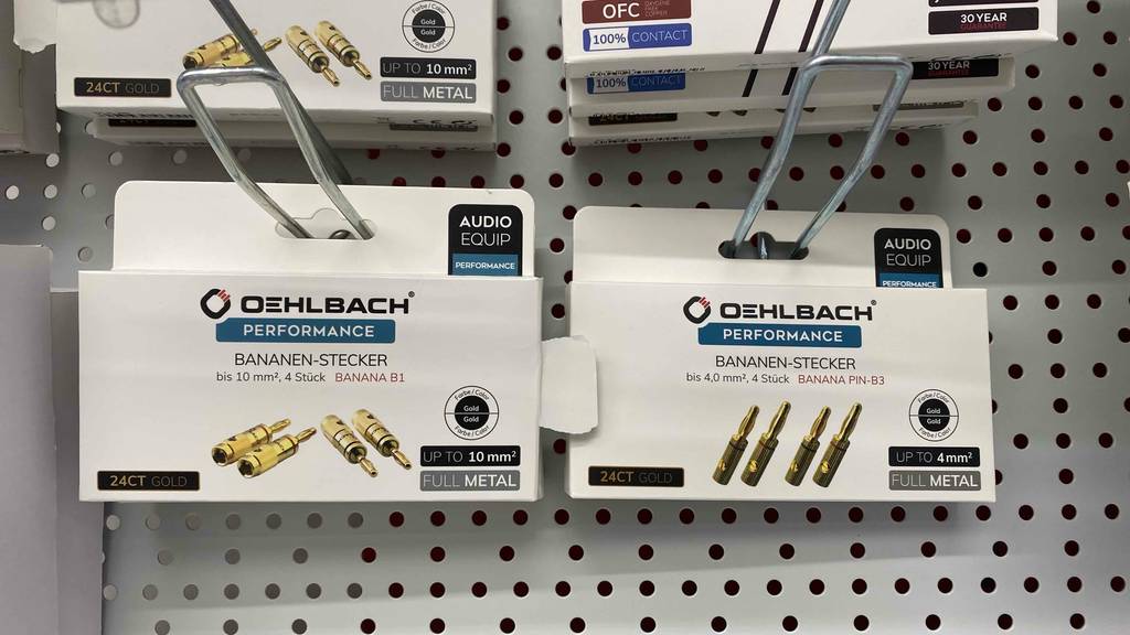 Mehrere unterschiedlich große Bananenstecker im Test: weiße Packungen inklusive vergoldeter Performance Bananen-Stecker von Oehlbach hängen an einem Verkaufsstand.