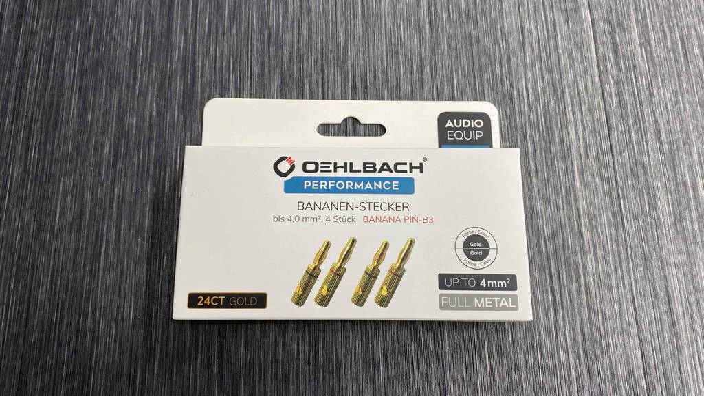 Bananenstecker im Test: eine weiße Packung inklusive vergoldeten Performance Bananen-Steckern von Oehlbach liegt auf einem grauen Untergrund.