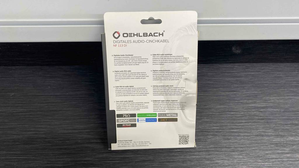 Ansicht der Rückseite inklusive Beschreibung einer weißen Verpackung von einem Oehlbach-Cinch-Kabel im Test, die auf einem grauen Untergrund steht.