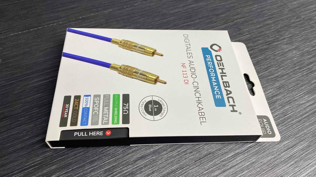 Die weiße Verpackung mit dem Oehlbach-Cinch-Kabel im Test inklusive goldener Stecker liegt auf einem grauen Boden.