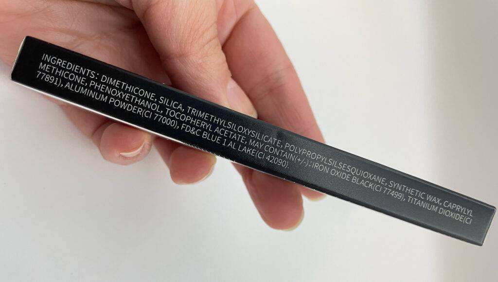 Eine Hand hält die Verpackung eines getesteten Gel-Eyeliners, mit Fokus auf die gedruckten Inhaltsstoffe über einer weißen Oberfläche.