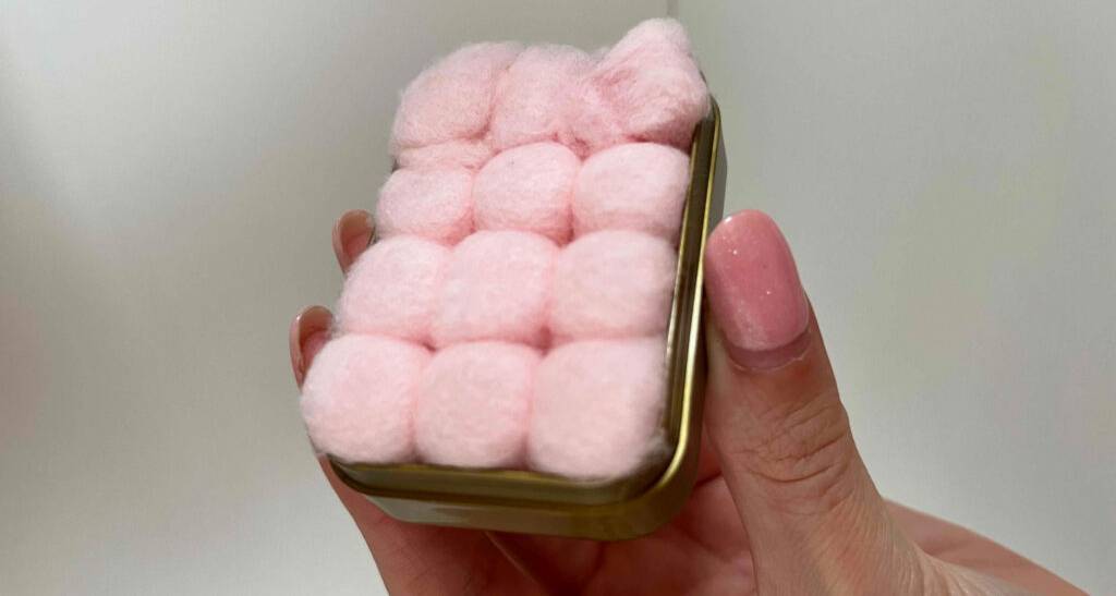 Damenfinger halten eine geöffnete Aufbewahrungsbox inklusive zwölf getesteter Konzert-Ohrstöpsel aus Wachs, mit rosa Watte umhüllt.