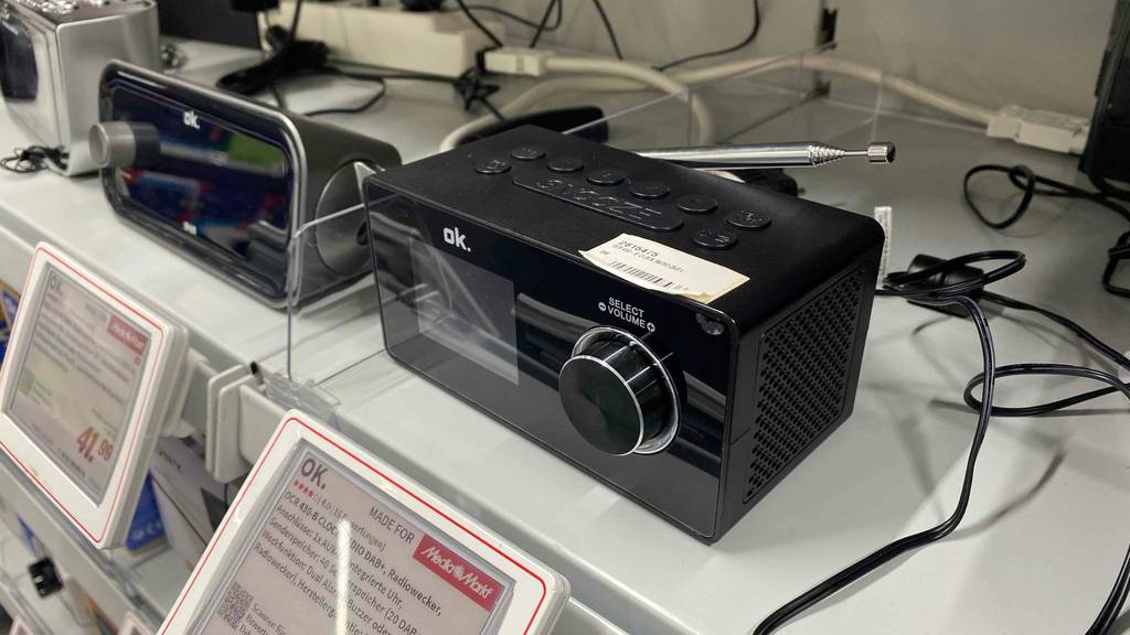 DAB-Radiowecker getestet: ein Modell in Schwarz der Marke ok. mit Digital-Anzeige und Drehknopf-Lautstärkeregler steht neben anderen elektronischen Geräten auf einem hellen Verkaufsregal.