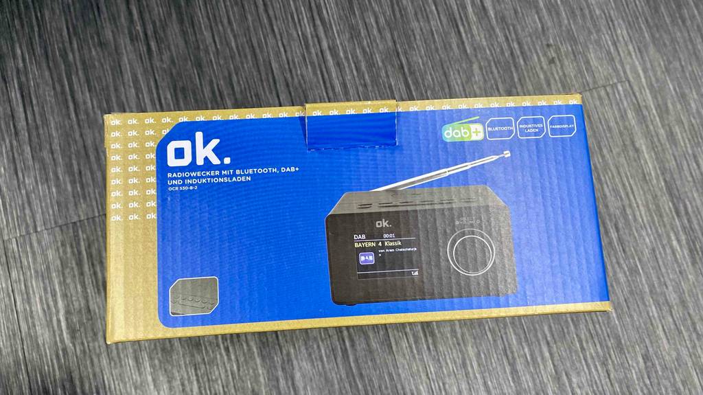 DAB-Radiowecker-Test: blau-goldene Verpackung eines Modells in Schwarz der Marke ok. mit Digital-Anzeige und Drehknopf-Lautstärkeregler liegt auf einem grauen Boden.