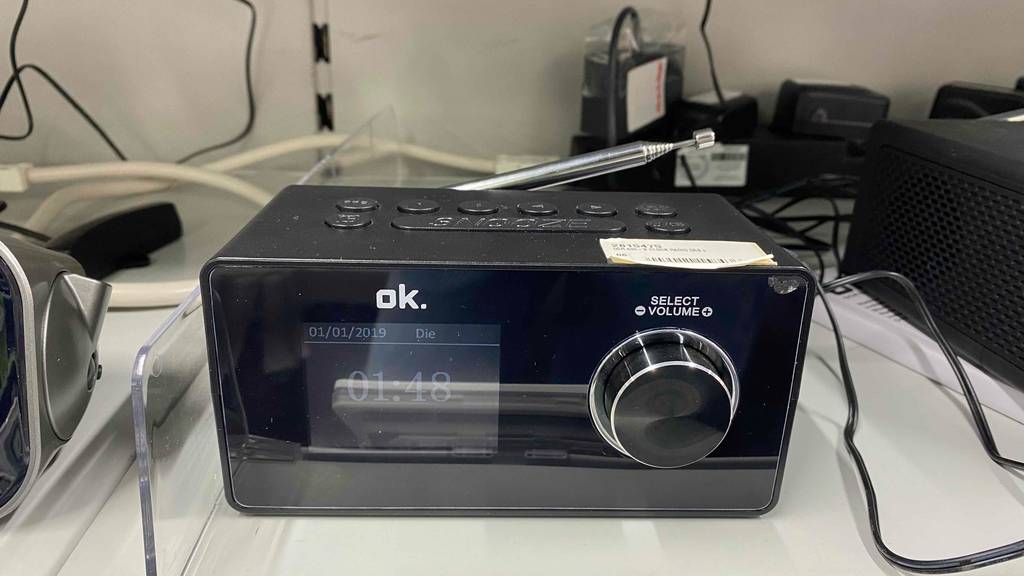 DAB-Radiowecker im Test: ein Modell in Schwarz der Marke ok. mit Digital-Anzeige und Drehknopf-Lautstärkeregler steht eingeschaltet auf einem hellen Verkaufsregal.