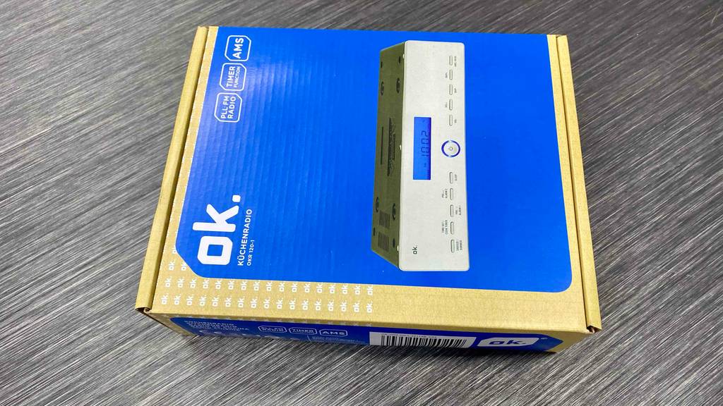 ok. Küchenradio getestet: Eine blau-goldene Verpackung eines AMS-Küchenradios inklusive Timer-Funktion liegt auf einem grauen Boden.