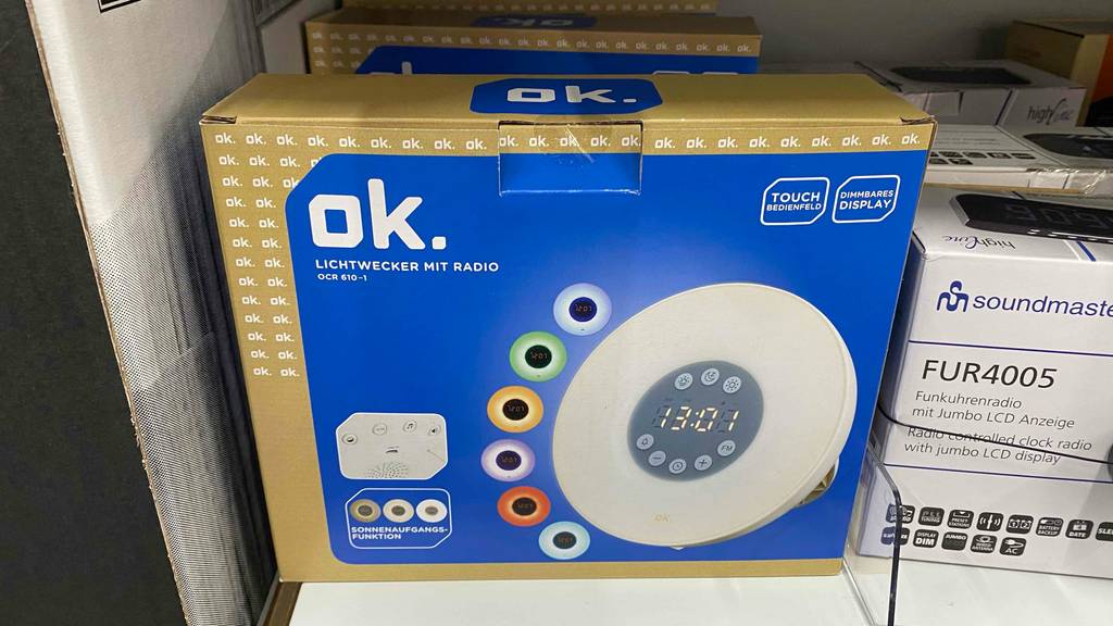 Eine blau-goldene Verpackung inklusive eines runden weißen ok. Lichtweckers im Test steht in einem Verkaufsregal sich auf einem grauen Untergrund.