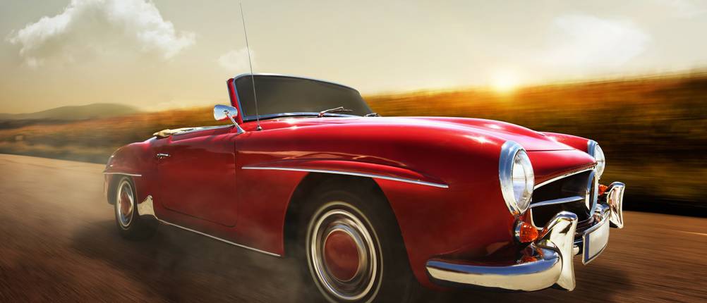 Rotes Oldtimer-Cabrio fährt mit Tempo auf einer Landstraße in der Abendsonne