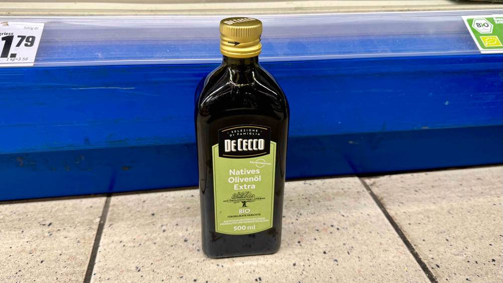 Getestet Olivenöl extra vergine: Eine Flasche vor einem Verkaufsregal mit Preisschildern am Boden stehend.